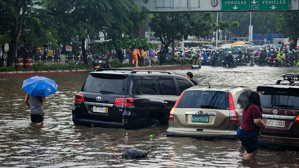 A flooded street with partially submerged cars, people wading through knee-deep water, and a person holding a blue umbrella. Crowds and bikes in the background convey chaos.