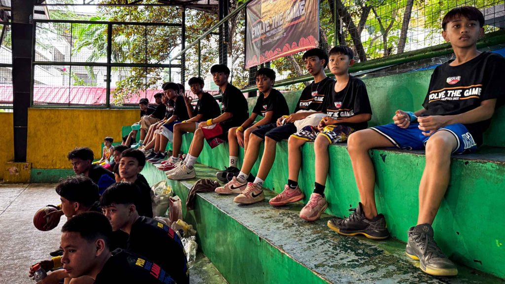 A group of boys in sportswear sit on green bleachers in an indoor basketball court. They look relaxed, holding basketballs, with trees visible outside.