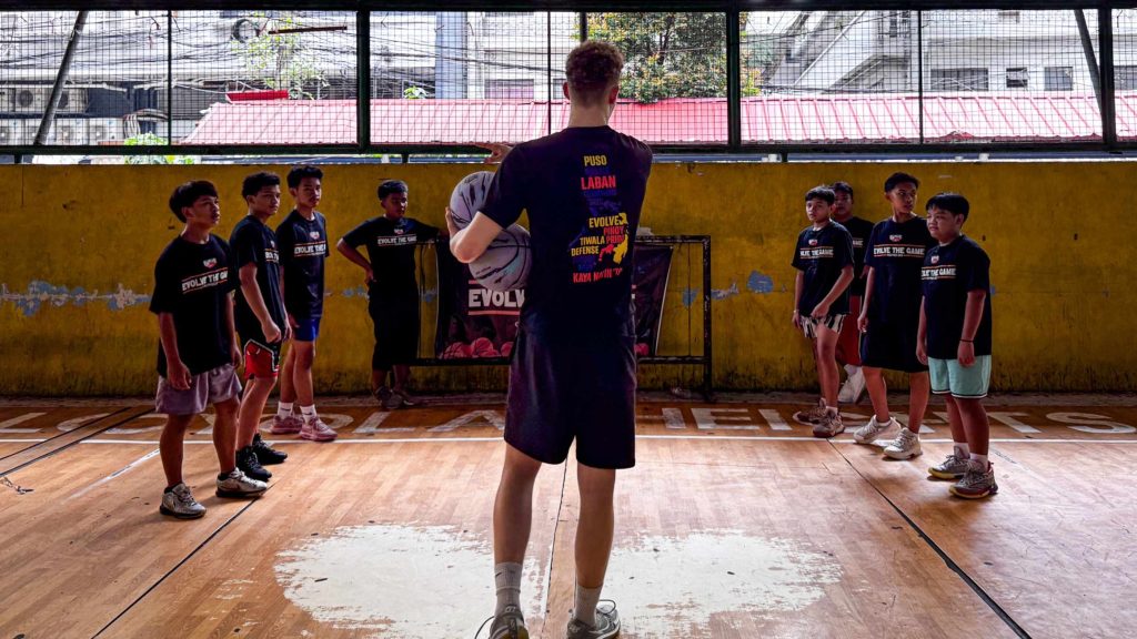 A coach with a basketball faces a group of young players on an indoor court, divided into two lines. The setting is informal and motivational.