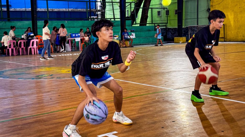 Two young men practice dribbling basketballs in an indoor gym. They wear black shirts and athletic gear, with a lively group in the background observing.