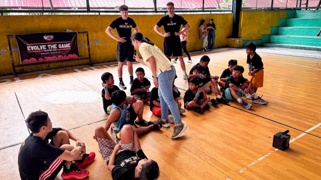 A group of young kids and a coach gather on a wooden basketball court. Kids sit on the floor holding basketballs, listening intently. Behind them stand three taller individuals in black shirts. The scene conveys teamwork and concentration. A banner on the wall reads "Evolve the Game."