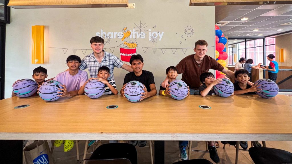 A group of children and two adults smile while seated at a table, each holding a signed basketball. The setting is festive, with colorful balloons in the background.