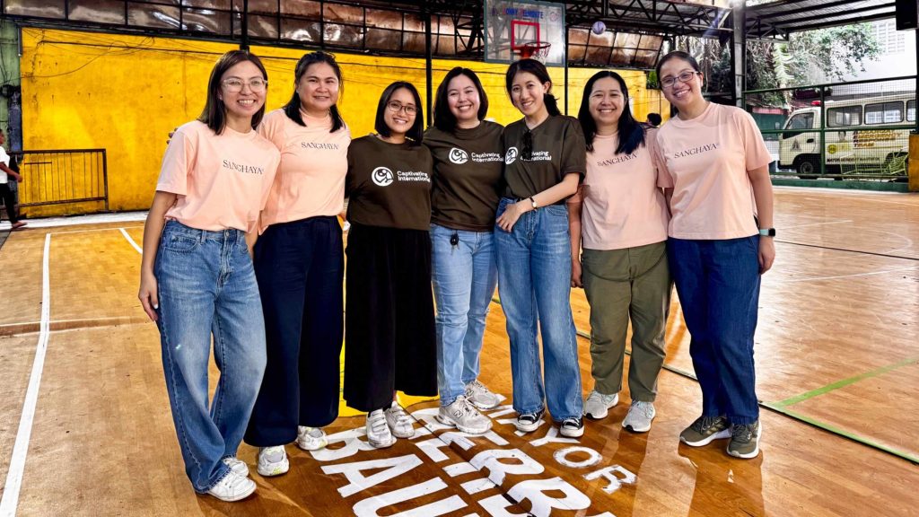 A group of seven smiling women poses indoors on a basketball court. They wear casual clothing in shades of blue and peach. The setting is bright and cheerful.