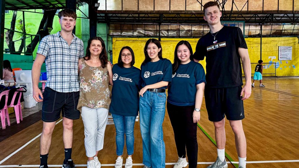 A group of six people smiling indoors on a wooden basketball court. The mood is friendly and relaxed.