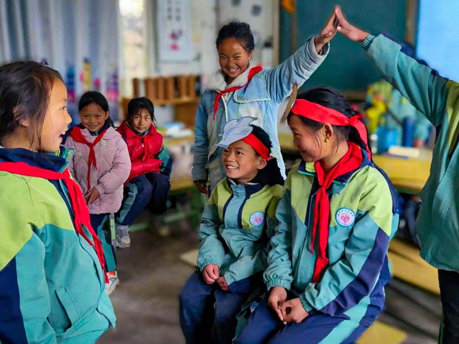 A group of smiling children in a classroom, wearing green jackets and red scarves. Two kids high-five, creating a joyful and playful atmosphere.