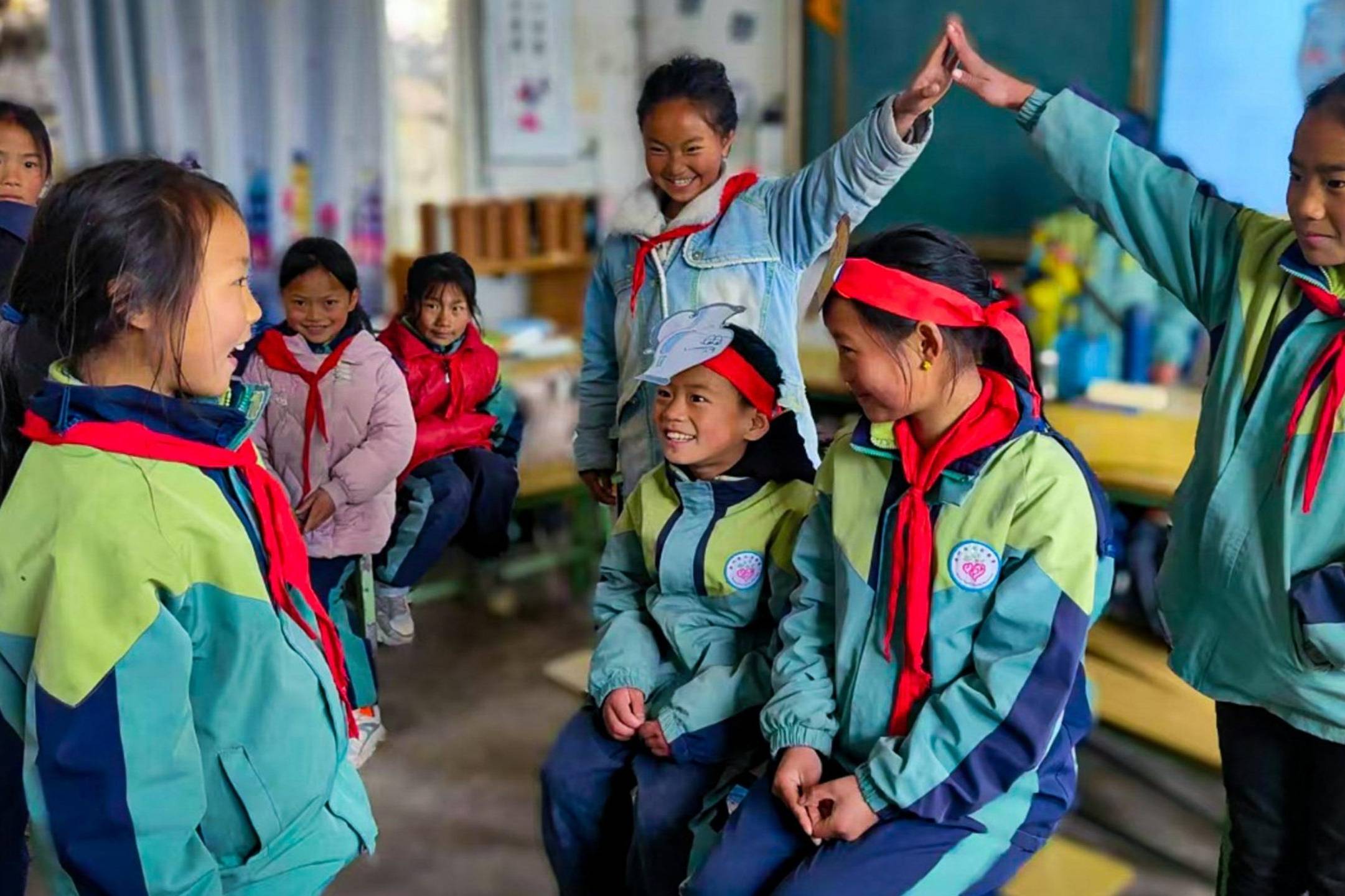 A group of smiling children in a classroom, wearing green jackets and red scarves. Two kids high-five, creating a joyful and playful atmosphere.