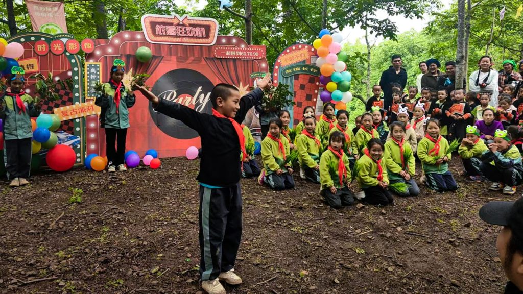 A joyful child in a red scarf performs with raised arms in front of an audience of children in matching green jackets. The outdoor scene is decorated with colorful balloons and a playful backdrop, evoking a festive atmosphere.