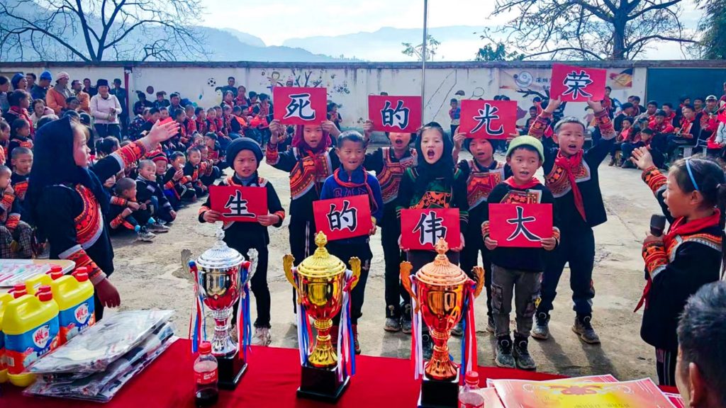 Children holding red cards with Chinese characters stand in a festive outdoor gathering. Three trophies are on a table, with a backdrop of trees and mountains.