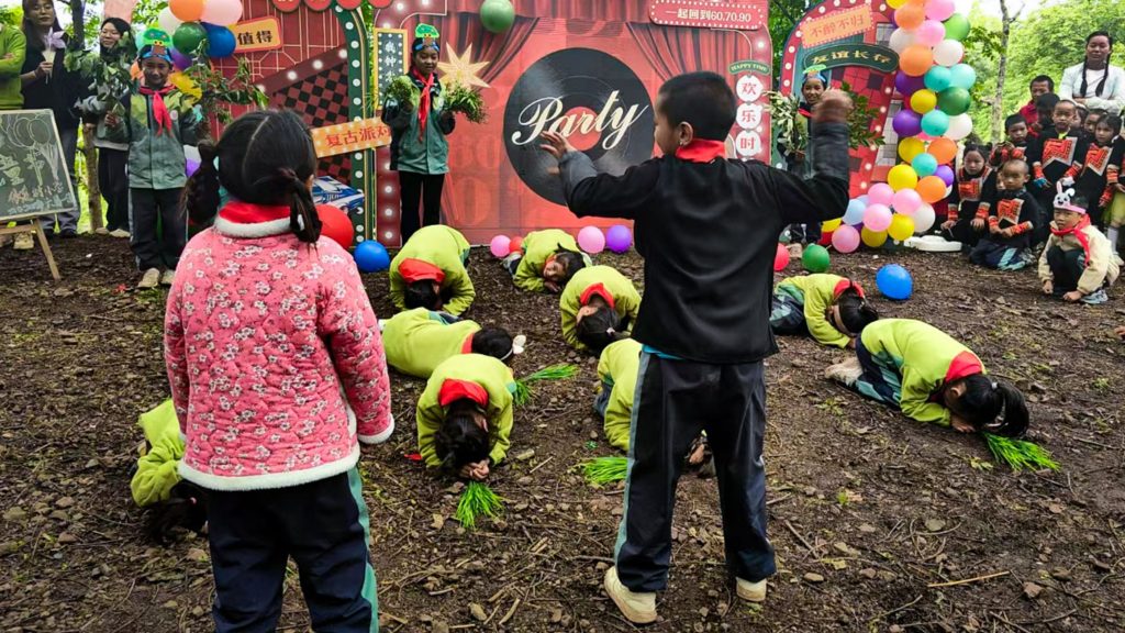 Children in colorful attire perform a coordinated dance on a dirt ground in front of a festive backdrop with balloons. The scene is lively and cheerful.