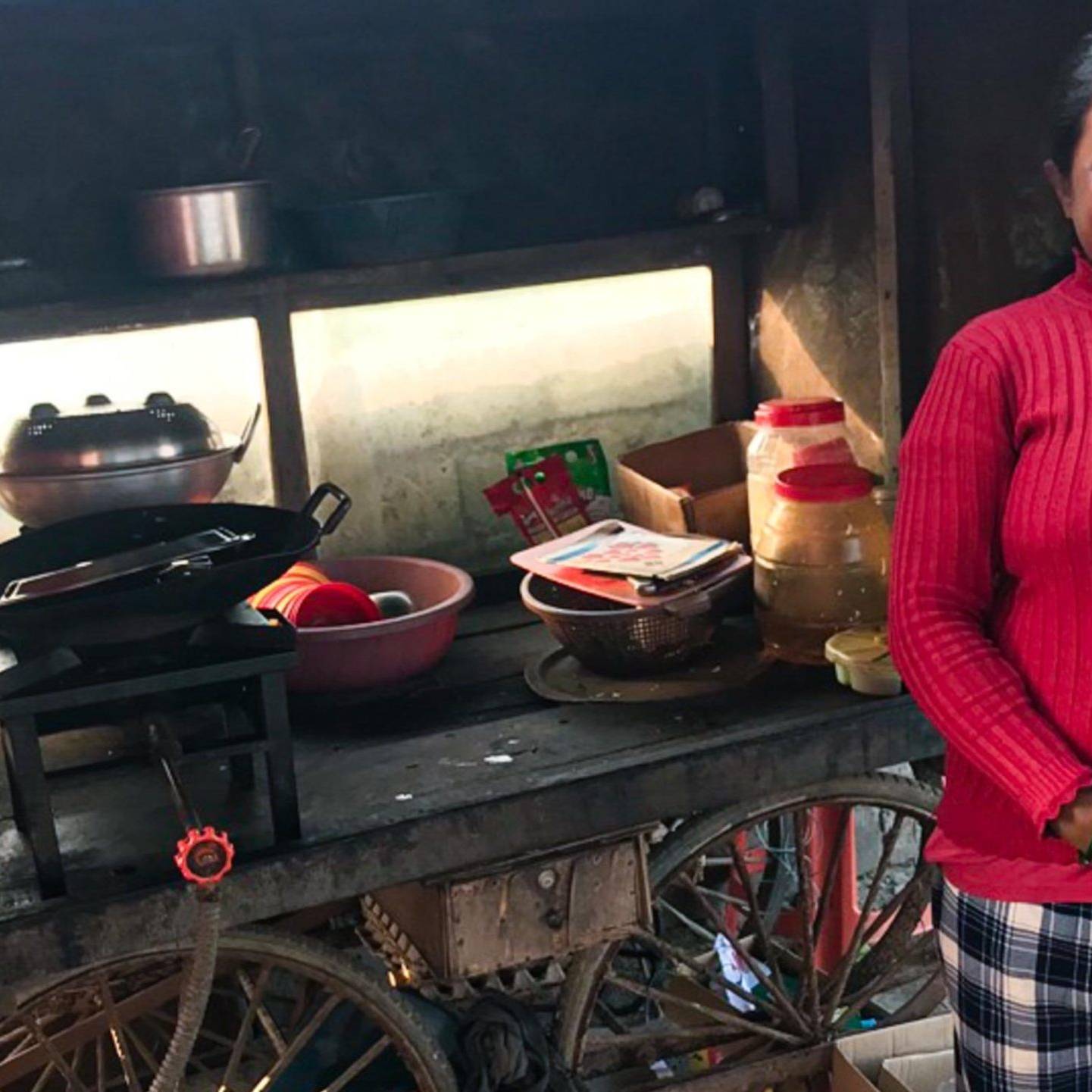 A woman in a red sweater stands smiling beside a wooden food cart. The cart has cooking pots, utensils, and jars, conveying a warm, welcoming vibe.