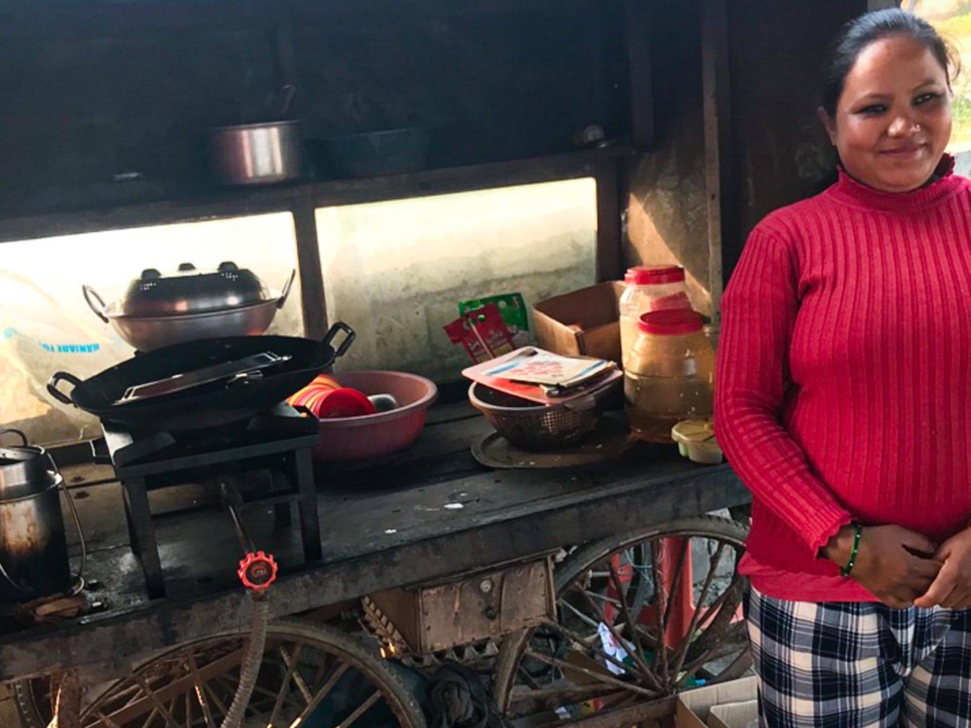 A woman in a red sweater stands smiling beside a wooden food cart. The cart has cooking pots, utensils, and jars, conveying a warm, welcoming vibe.