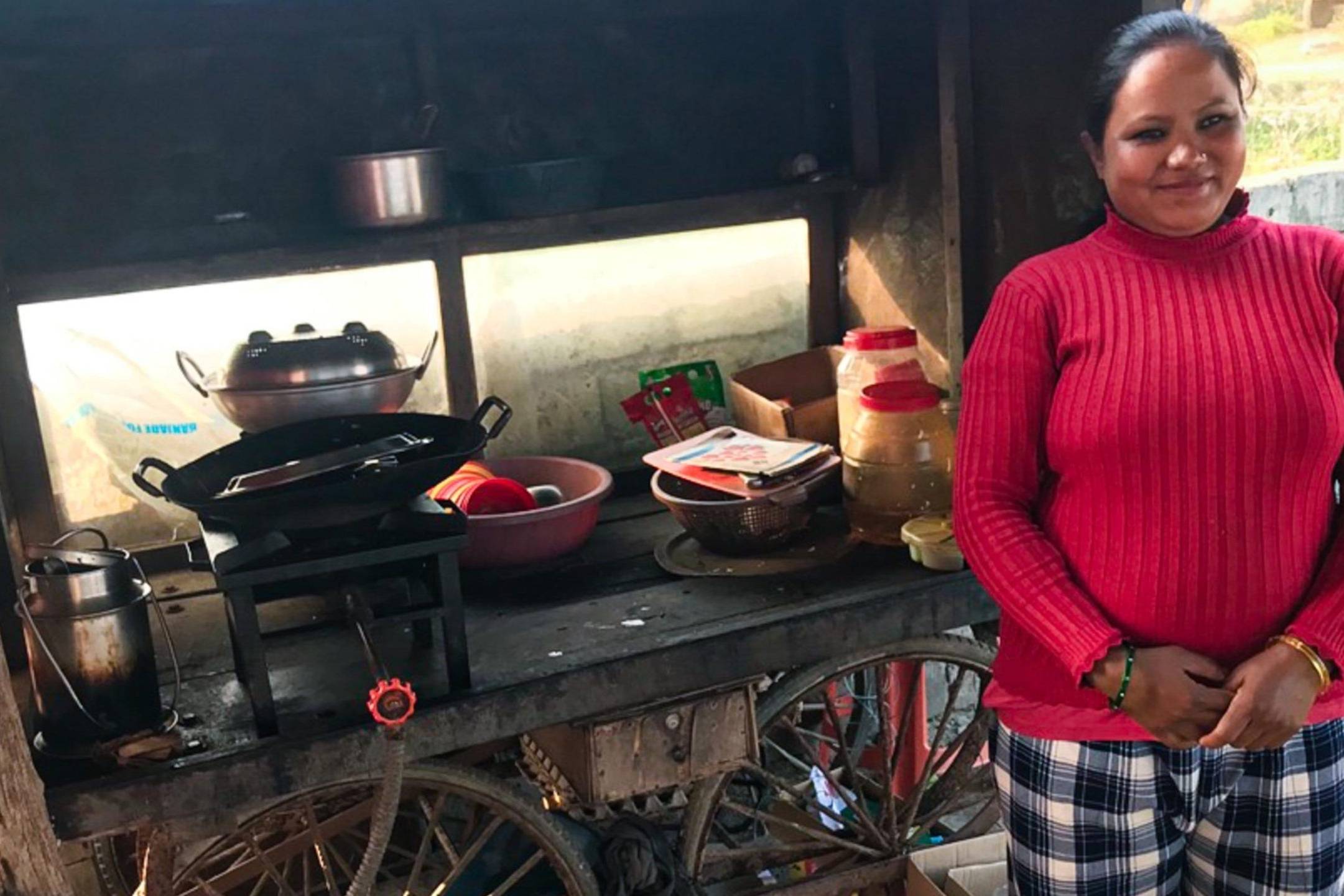 A woman in a red sweater stands smiling beside a wooden food cart. The cart has cooking pots, utensils, and jars, conveying a warm, welcoming vibe.