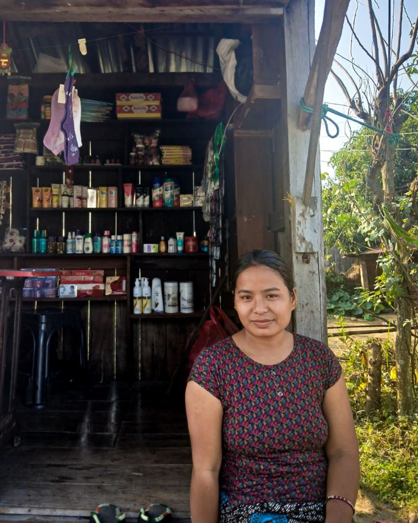 A woman stands outside a small wooden shop filled with neatly arranged toiletries and household items.