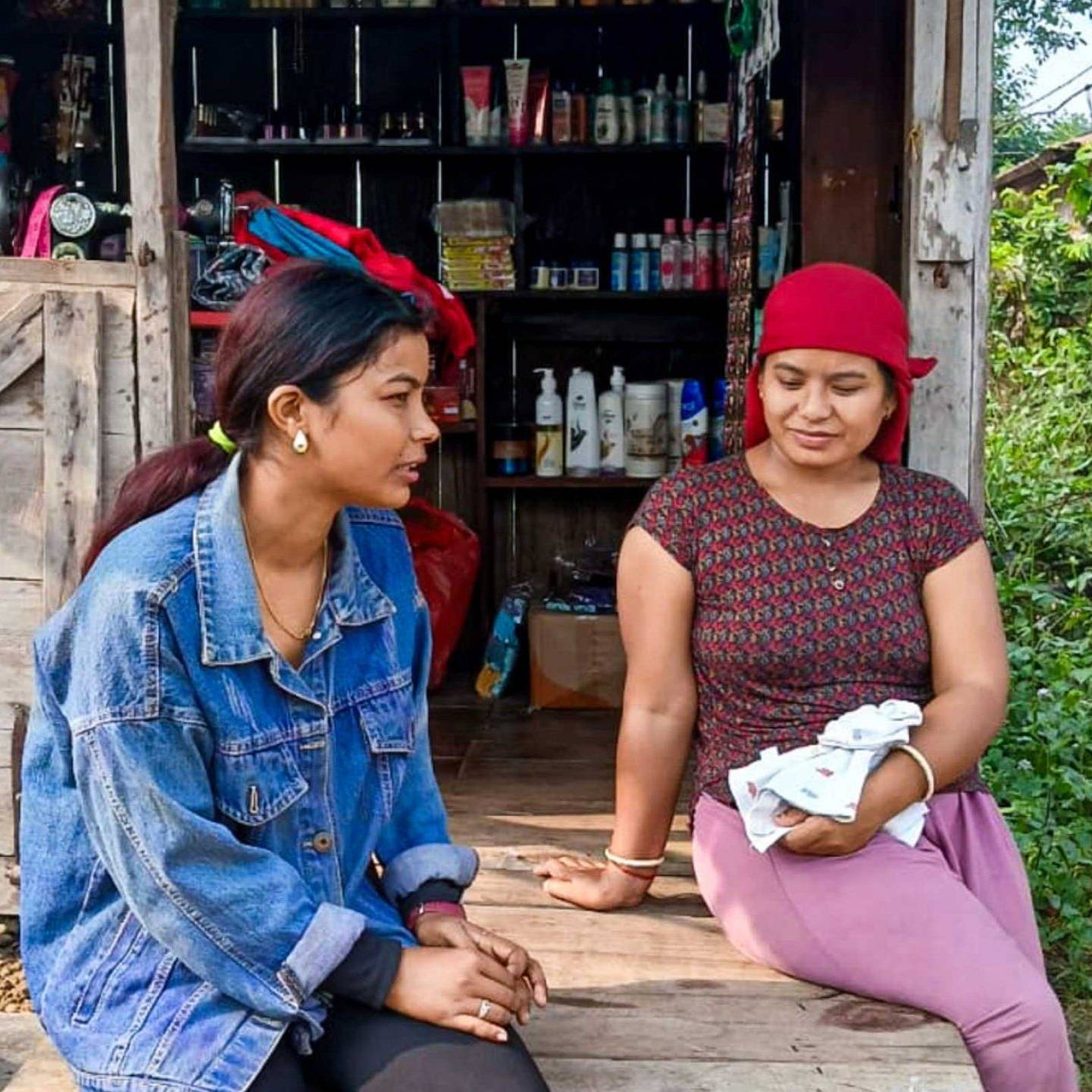 Two women sit on a wooden bench outside a small shop with various items. One wears a denim jacket; the other cradles a baby, exuding a relaxed, rustic mood.
