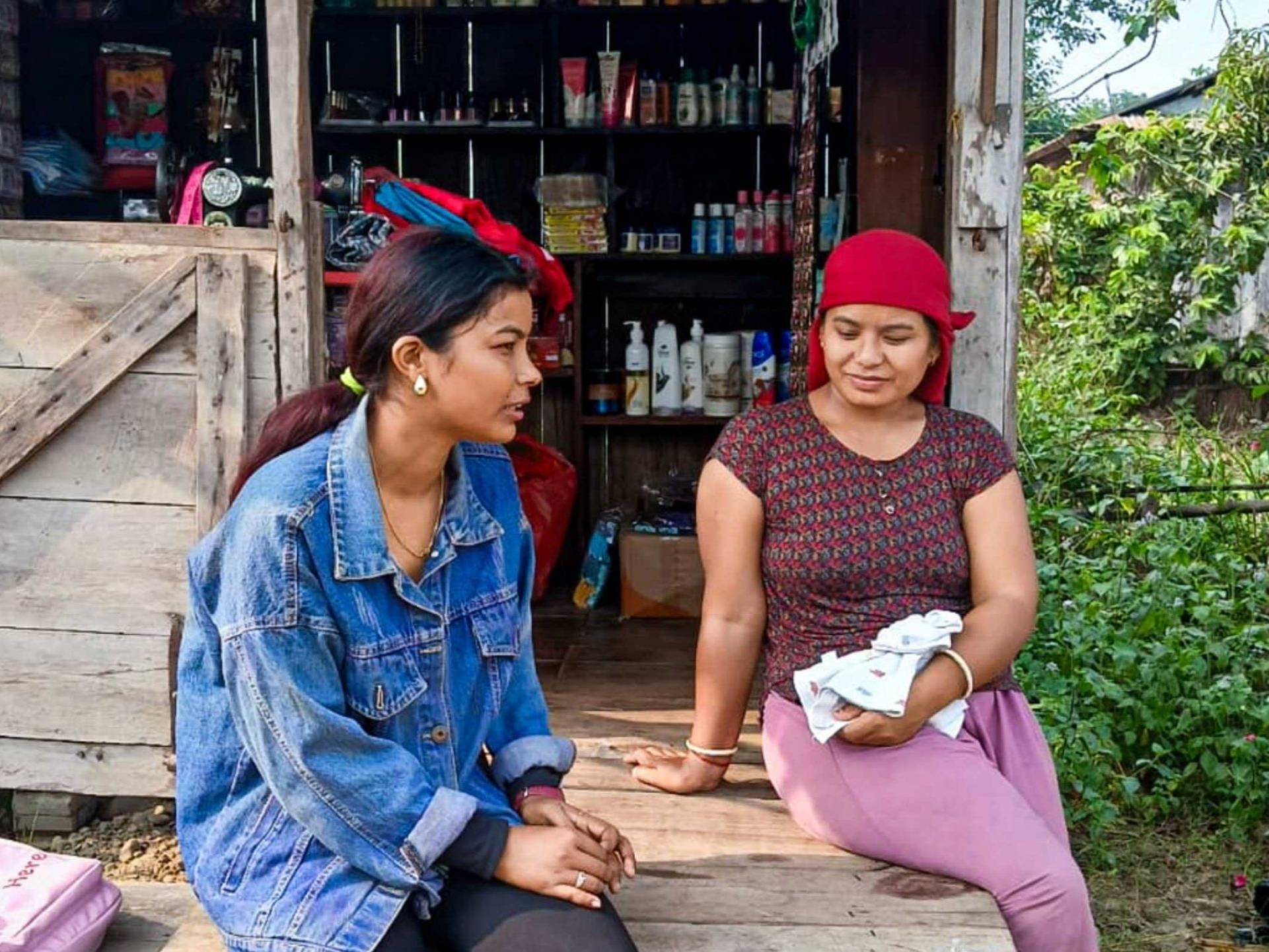 Two women sit on a wooden bench outside a small shop with various items. One wears a denim jacket; the other cradles a baby, exuding a relaxed, rustic mood.