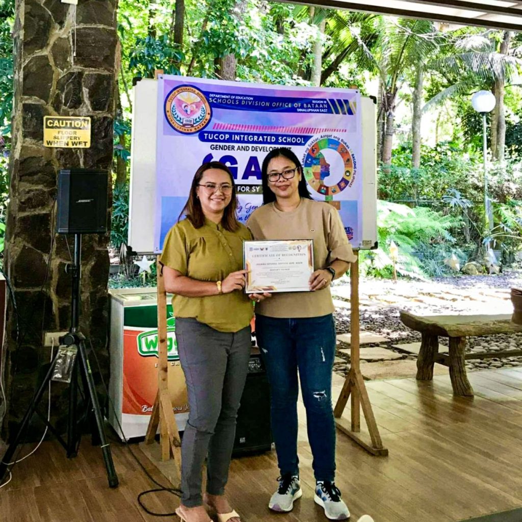 Two people smiling while holding a certificate in an outdoor setting with tropical plants. A banner in the background suggests a school event.