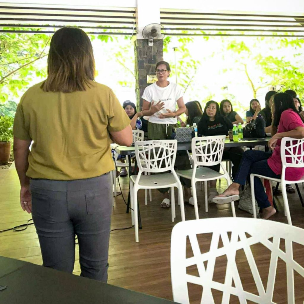 A woman in a white shirt stands speaking to a group seated around tables indoors. The setting is bright with greenery outside, suggesting a calm, focused meeting.