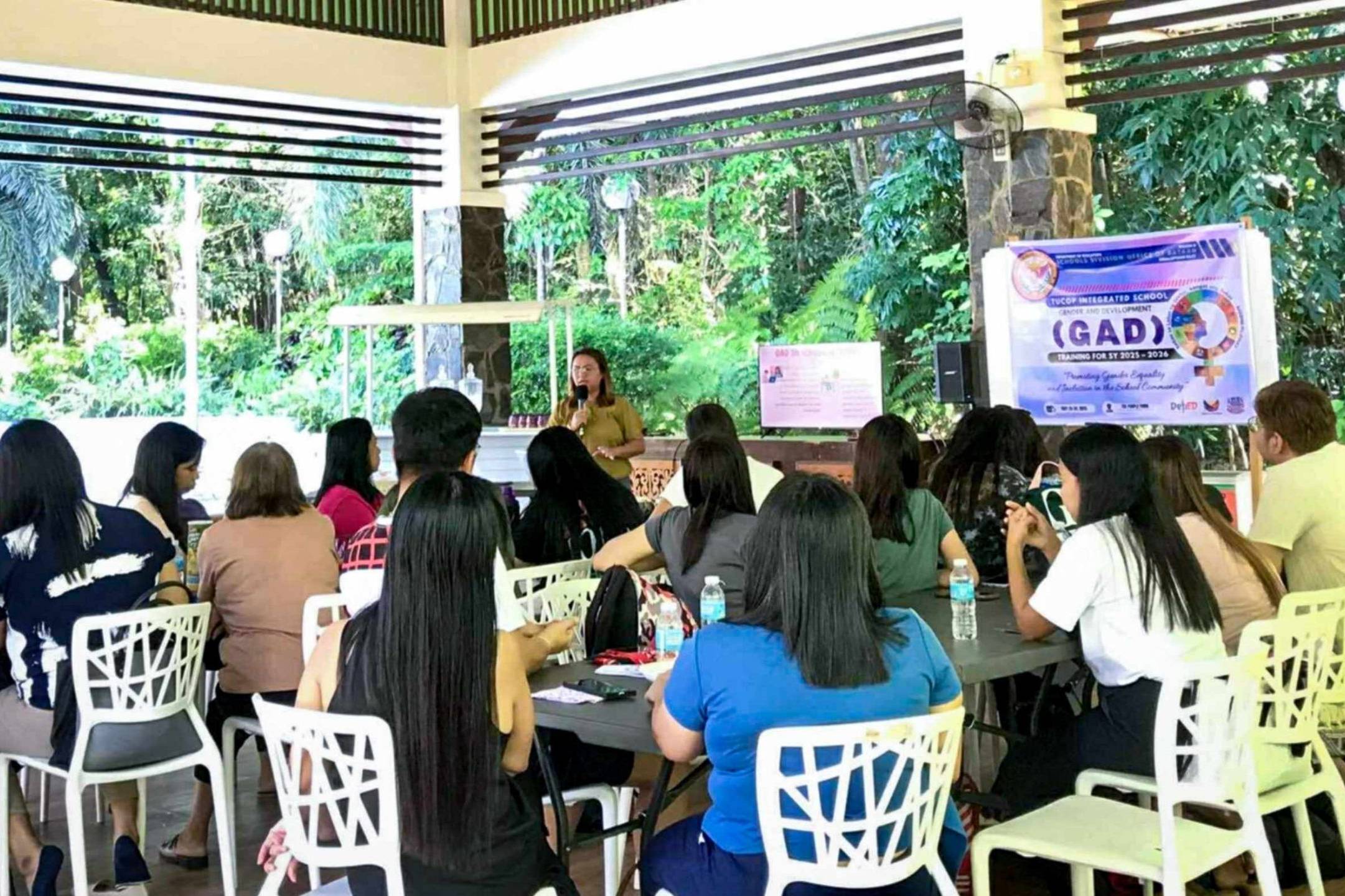 A woman speaks to a seated audience in an airy pavilion with a peaked roof. Attendees listen intently.