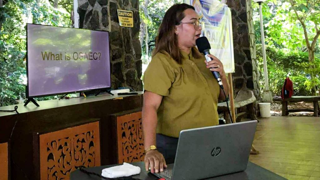 A woman in a mustard blouse speaks into a microphone during a presentation.