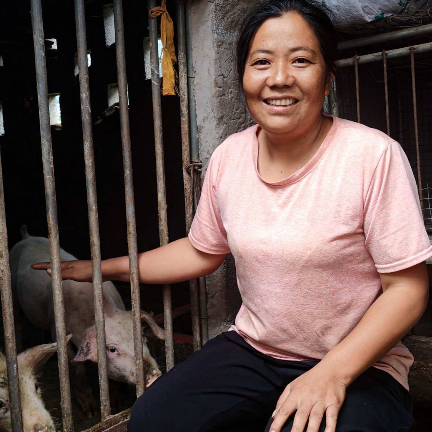 A woman in a pink shirt smiles warmly, sitting beside a pigpen with pigs inside.