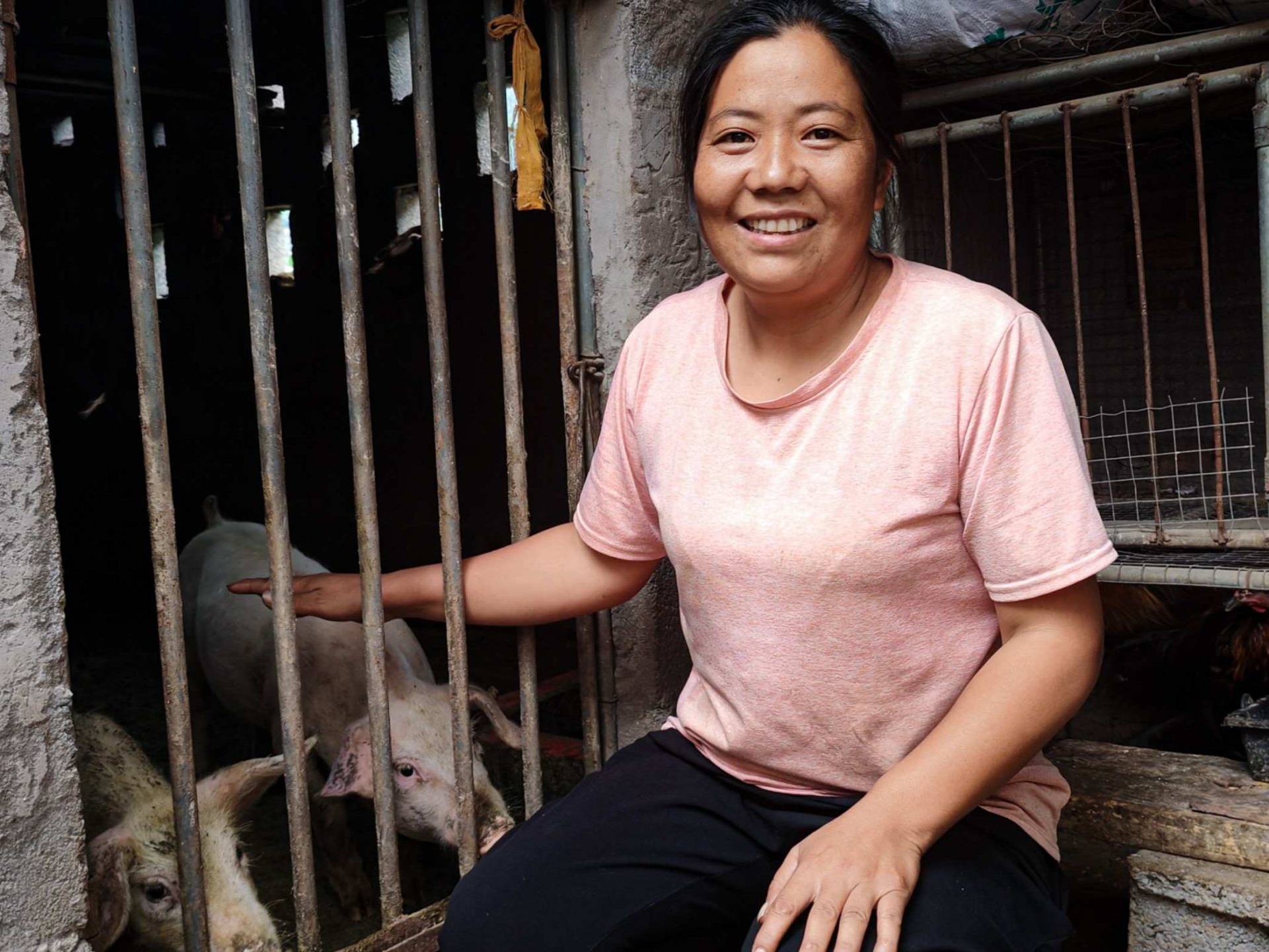 A woman in a pink shirt smiles warmly, sitting beside a pigpen with pigs inside.