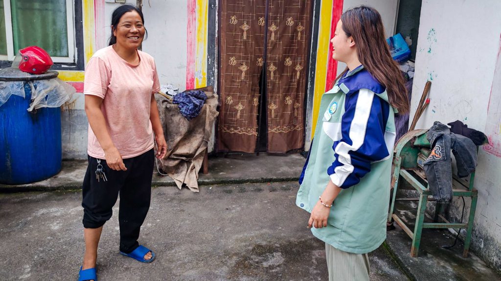 Two women are smiling at each other outside a building. One wears a pink shirt; the other, a blue and green jacket.