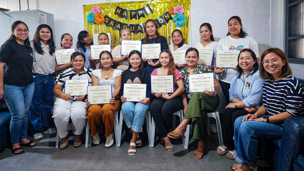 Group of women smiling and holding graduation certificates, sitting in two rows. Behind them is a “Happy Graduation” banner with colorful decorations.