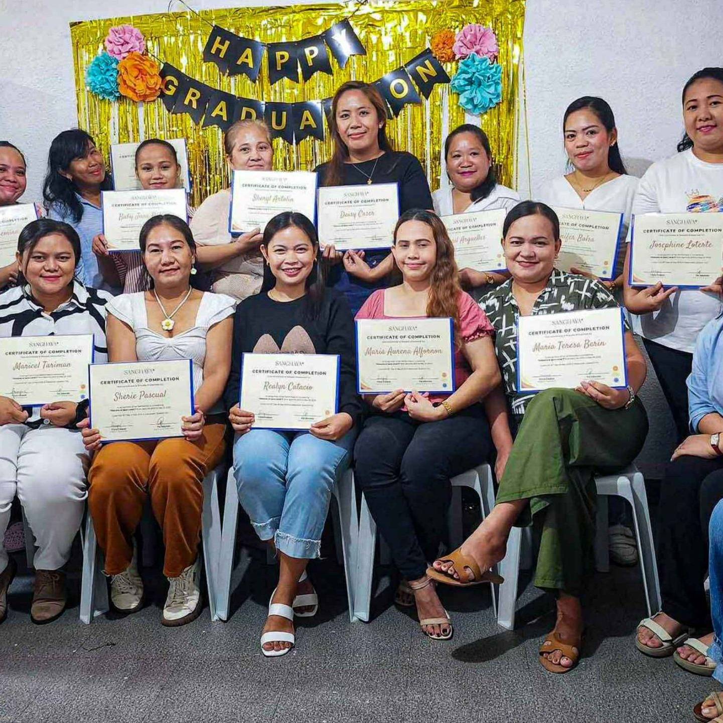 Group of women smiling and holding graduation certificates, sitting in two rows. Behind them is a “Happy Graduation” banner with colorful decorations.