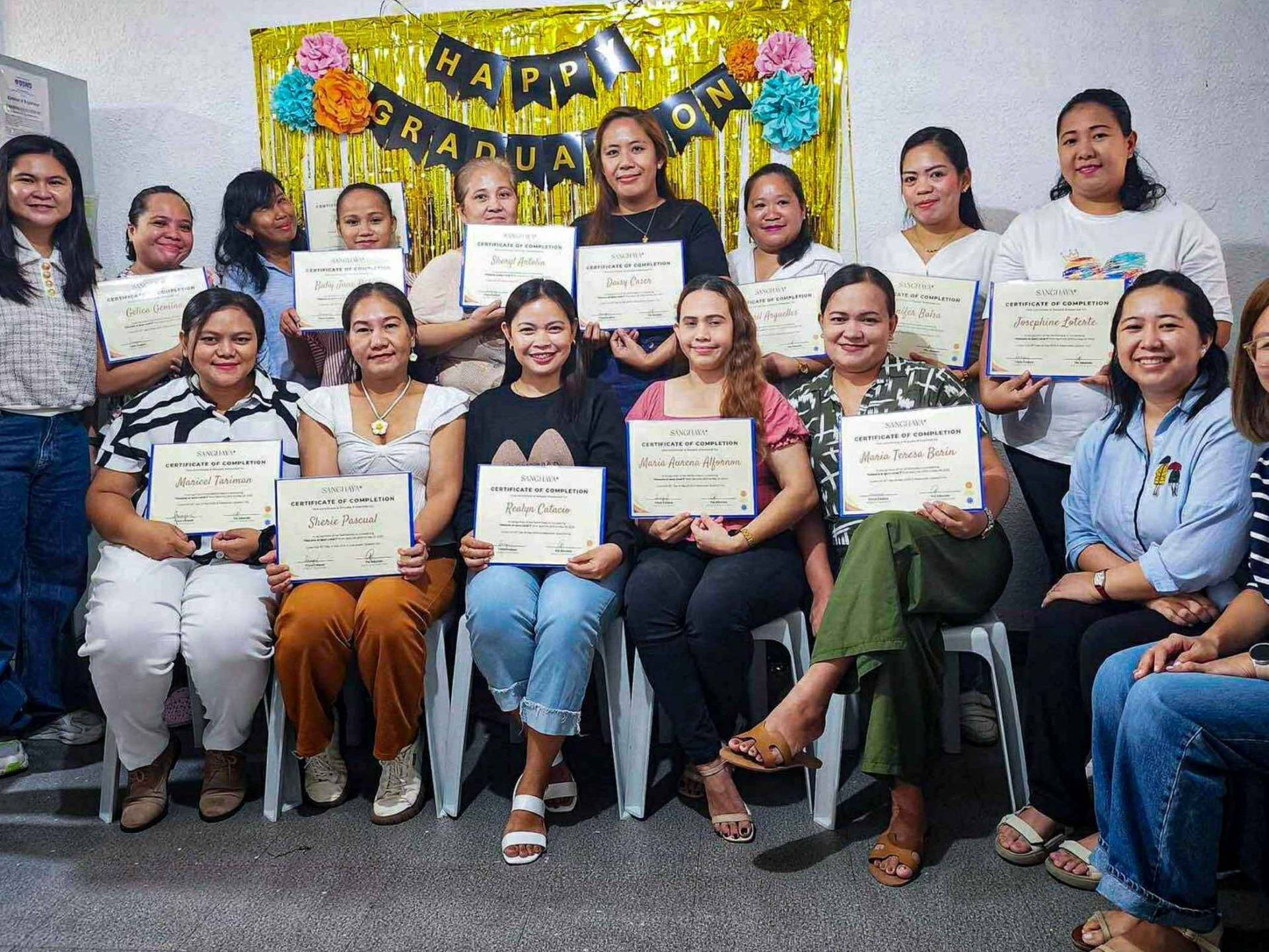 Group of women smiling and holding graduation certificates, sitting in two rows. Behind them is a “Happy Graduation” banner with colorful decorations.