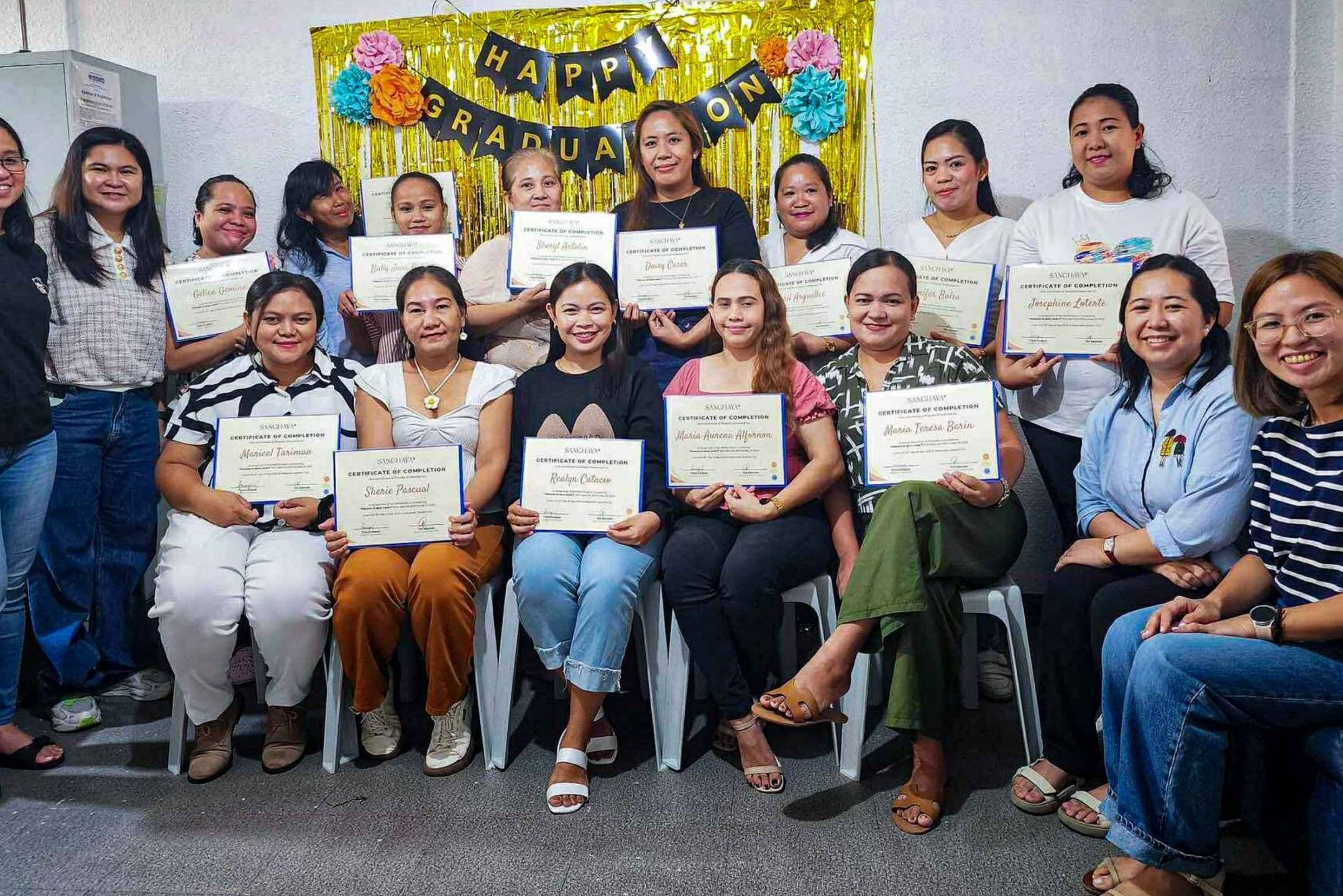 Group of women smiling and holding graduation certificates, sitting in two rows. Behind them is a “Happy Graduation” banner with colorful decorations.