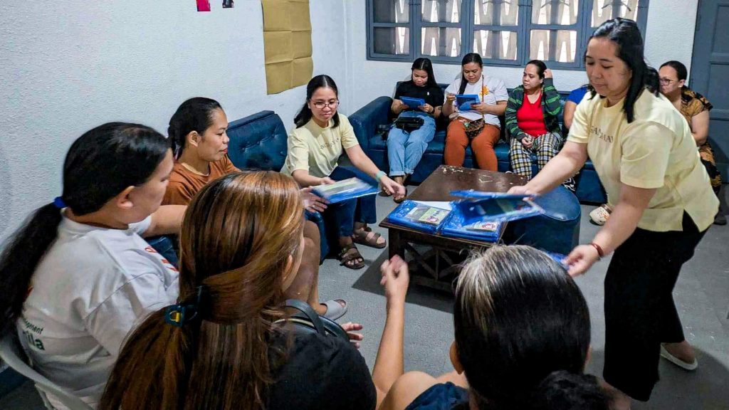 A group of women sits in a cozy room, some holding documents. The atmosphere is collaborative and focused.