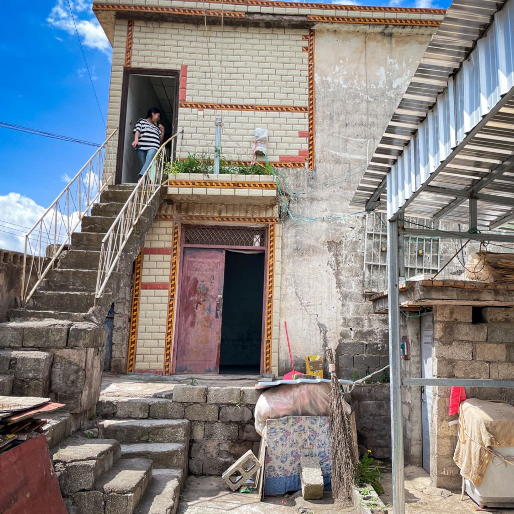 A two-story brick house with a person standing on the outdoor staircase. The yard is cluttered with stone, metal, and covered furniture. Bright, sunny day.