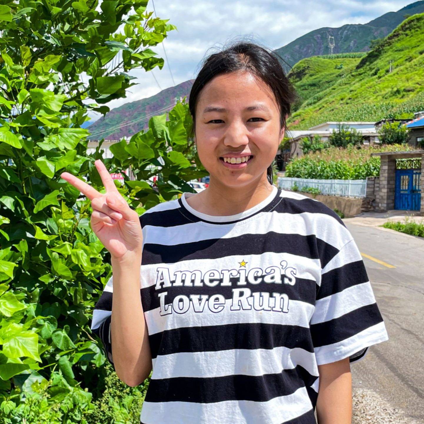 A smiling person in a striped shirt shows a peace sign. They stand by lush greenery on a sunny day, with a rural road and hills in the background.