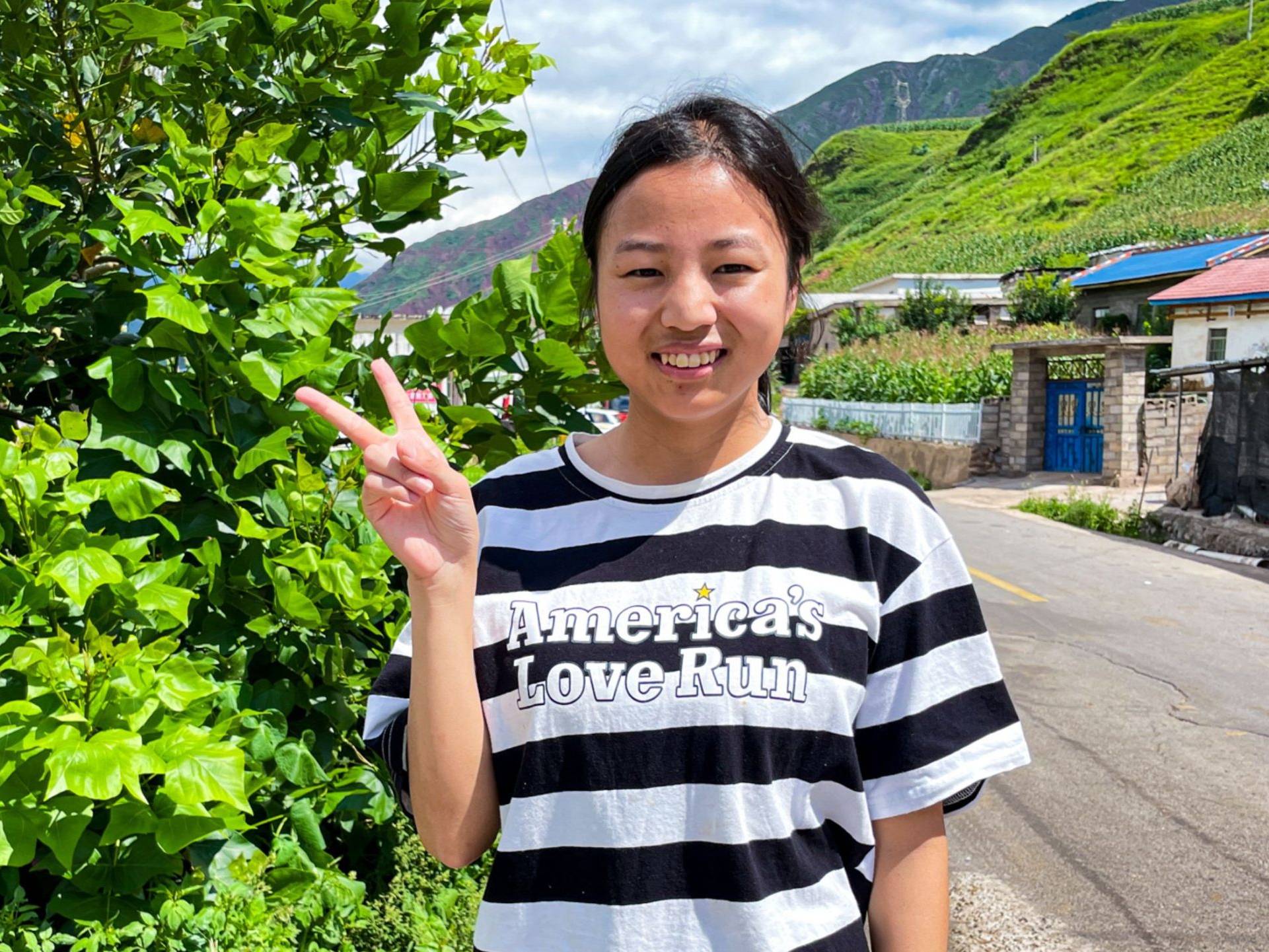 A smiling person in a striped shirt shows a peace sign. They stand by lush greenery on a sunny day, with a rural road and hills in the background.