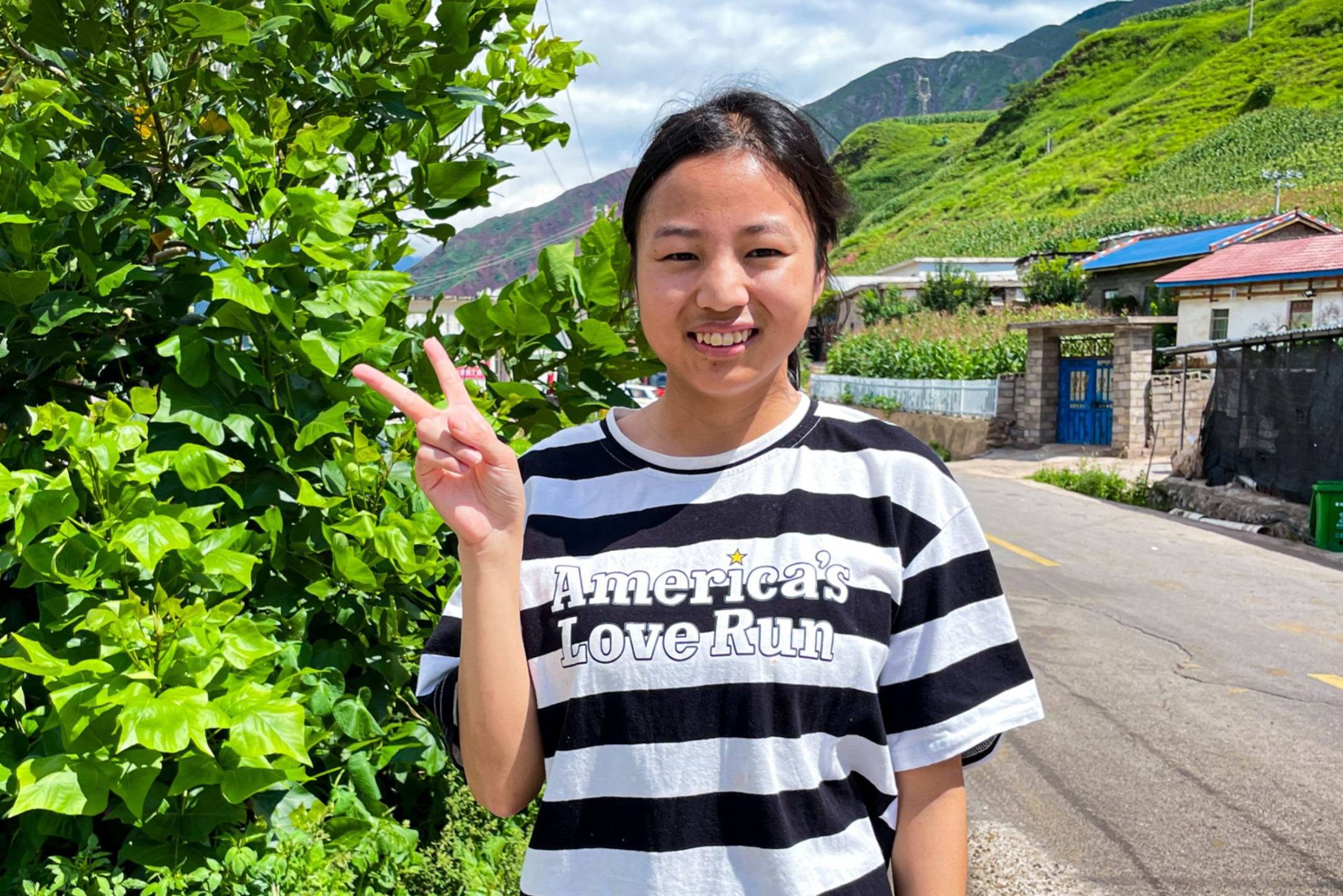 A smiling person in a striped shirt shows a peace sign. They stand by lush greenery on a sunny day, with a rural road and hills in the background.
