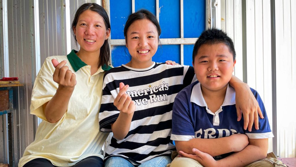 A joyful group of three, including two women and a boy, sit closely on steps. They smile warmly, making small heart gestures with their fingers, creating a sense of unity.