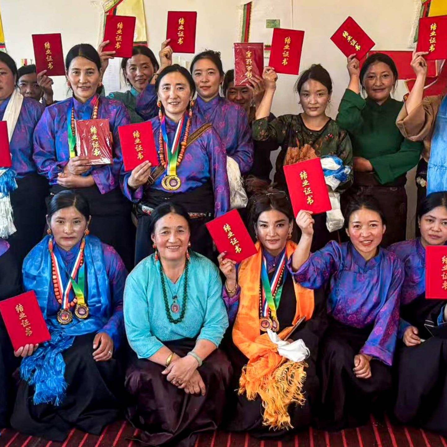 A group of women in vibrant blue and purple traditional attire hold red certificates proudly, smiling warmly. The setting is a festive indoor space.