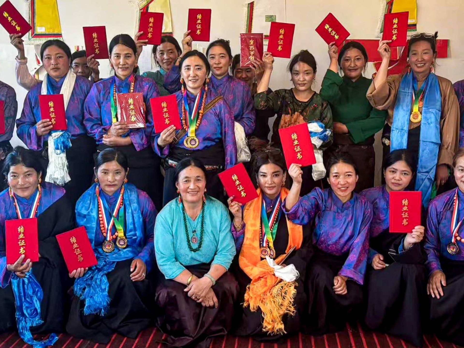 A group of women in vibrant blue and purple traditional attire hold red certificates proudly, smiling warmly. The setting is a festive indoor space.