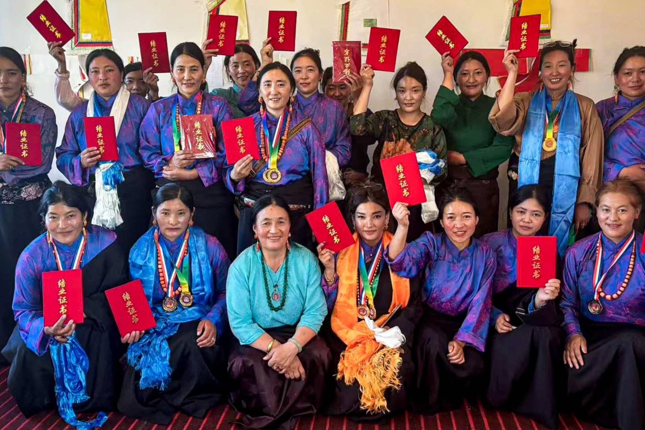 A group of women in vibrant blue and purple traditional attire hold red certificates proudly, smiling warmly. The setting is a festive indoor space.