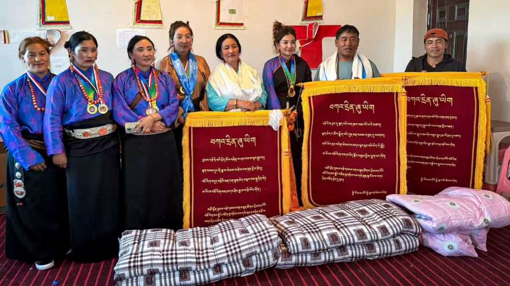 A group of eight people stands indoors, wearing traditional attire with medals and scarves. They pose in front of red banners with Tibetan script, exuding a celebratory and proud atmosphere. In front of them are folded plaid and floral blankets.