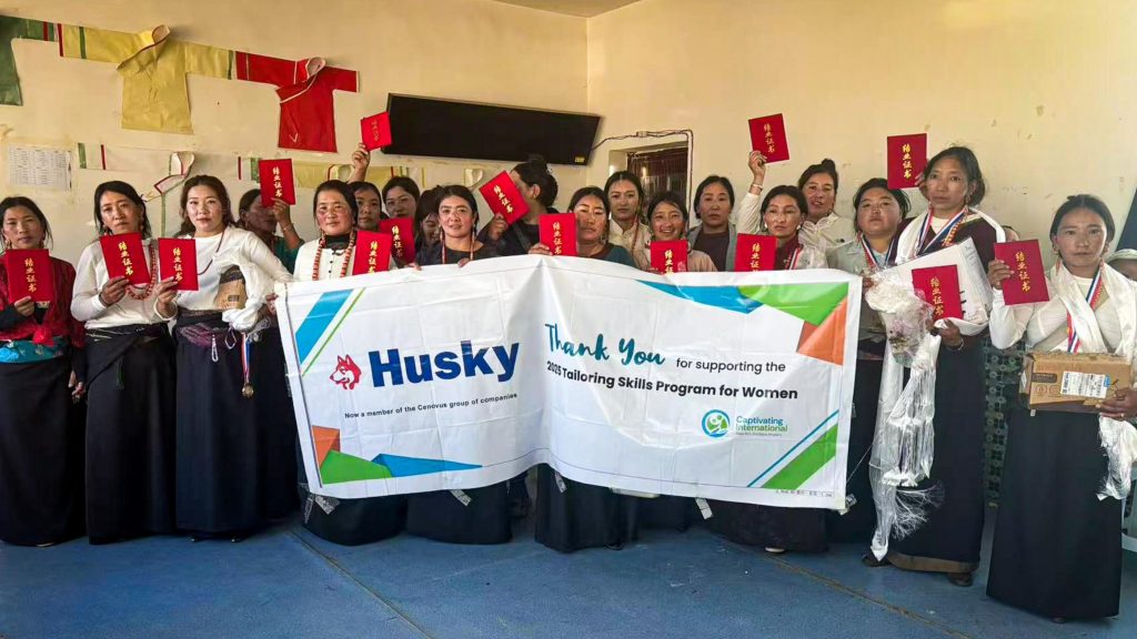 A group of women stands indoors, holding red certificates and medals, smiling proudly. A banner in front thanks supporters of a women's tailoring skills program.