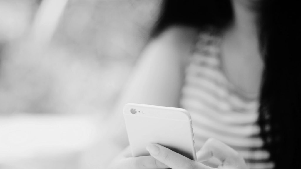 A person holding a smartphone with focus on the device. The background is blurred, creating a relaxed and casual mood. The image is in black and white.
