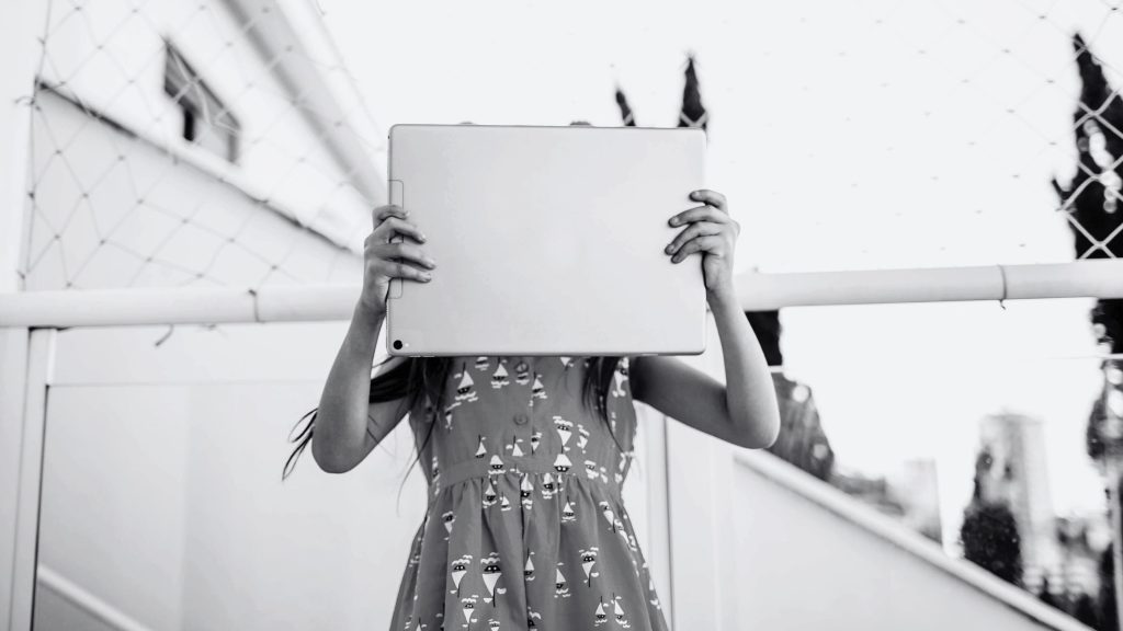 Child in a patterned dress holds a laptop covering their face, standing outdoors near a netted fence. The scene conveys a playful, mysterious mood.