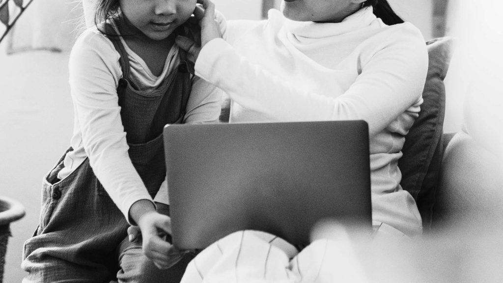 A woman and a young girl sit together on a couch, engaged with a laptop. The woman gently holds the girl's shoulder, conveying a warm, nurturing atmosphere.