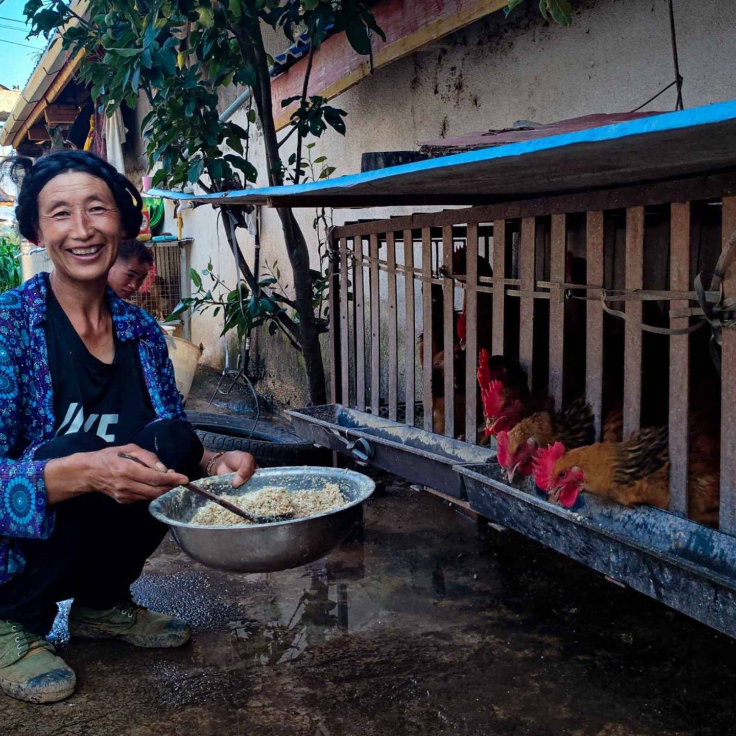 A woman smiles facing the camera while crouching beside a hen coop, holding a bowl of feed. Chickens eat from the coop. The scene conveys warmth and rural life.