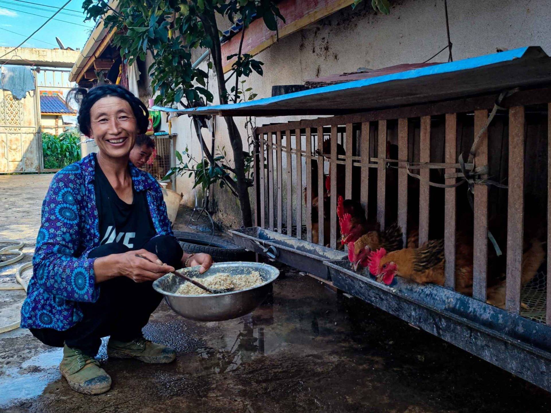 A woman smiles facing the camera while crouching beside a hen coop, holding a bowl of feed. Chickens eat from the coop. The scene conveys warmth and rural life.