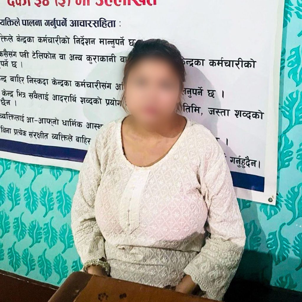 A person in a white patterned blouse is seated in front of a wall with Nepali text. Face blurred for privacy. The wall is teal with a leaf design.