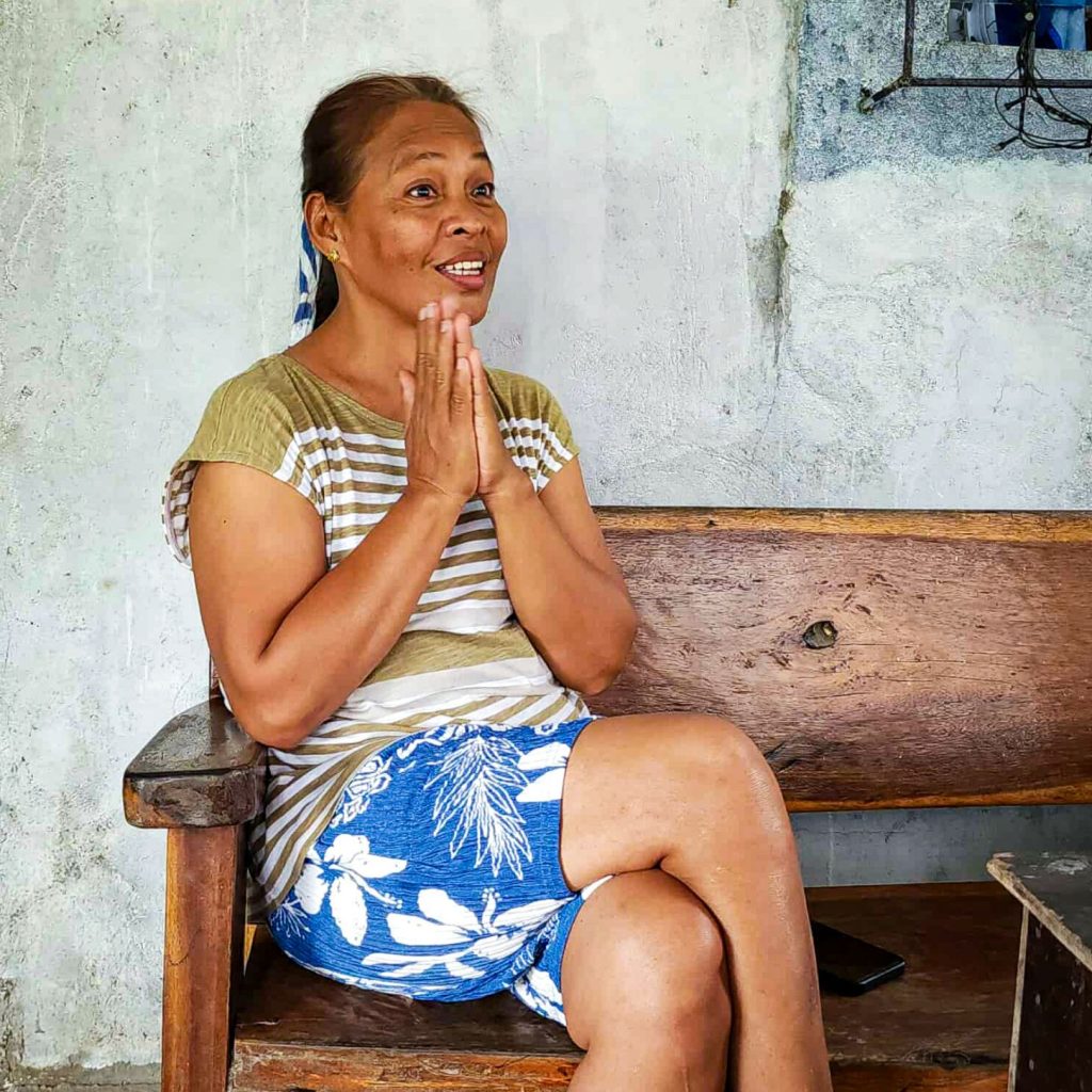 A woman sits on a wooden bench, smiling with hands clasped in front. She's wearing a striped shirt and floral shorts, conveying warmth and joy.