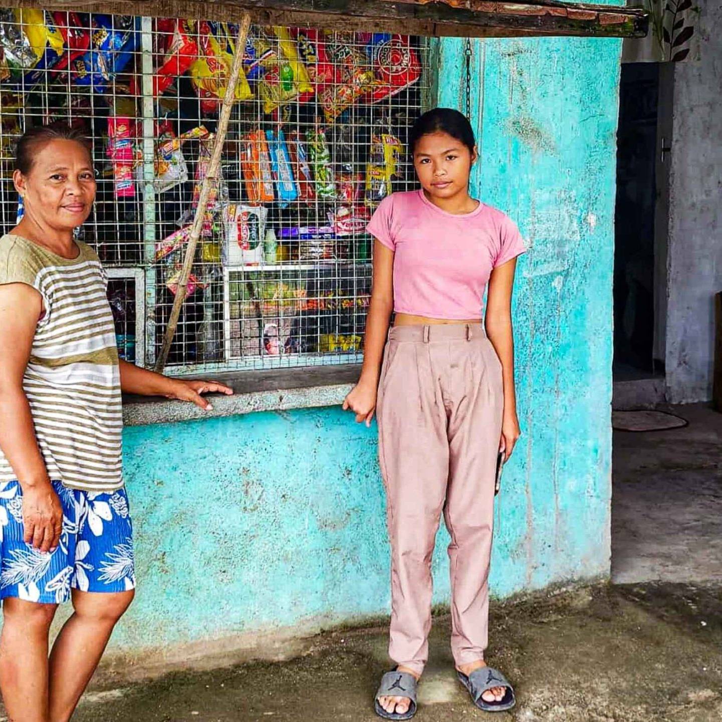Two people stand outside a small shop with teal walls and a metal grille. The woman is in striped shirt and shorts; the girl wears a pink shirt and beige pants. The shop is stocked with snacks, creating a warm community vibe.