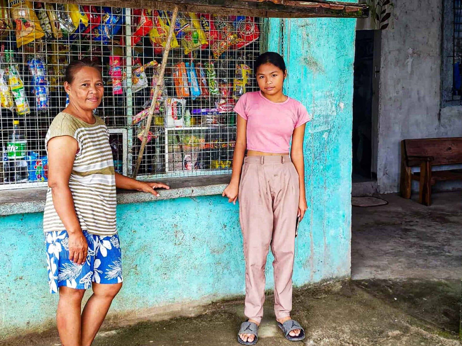 Two people stand outside a small shop with teal walls and a metal grille. The woman is in striped shirt and shorts; the girl wears a pink shirt and beige pants. The shop is stocked with snacks, creating a warm community vibe.